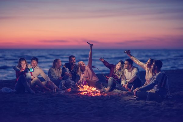 Group of young couples taking selfies by the fire on the beach at sunset