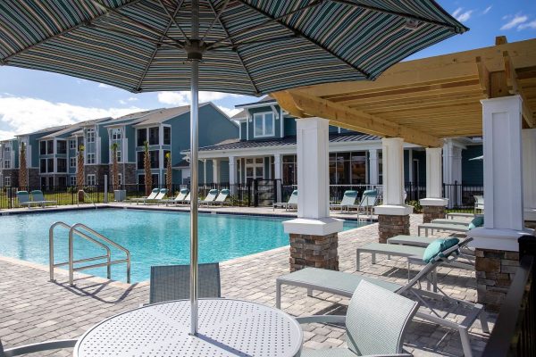 Bright blue, resort-style swimming pool with expansive sundeck and ample lounging options including shaded areas, all fenced-in in front of the clubhouse and some apartment homes.
